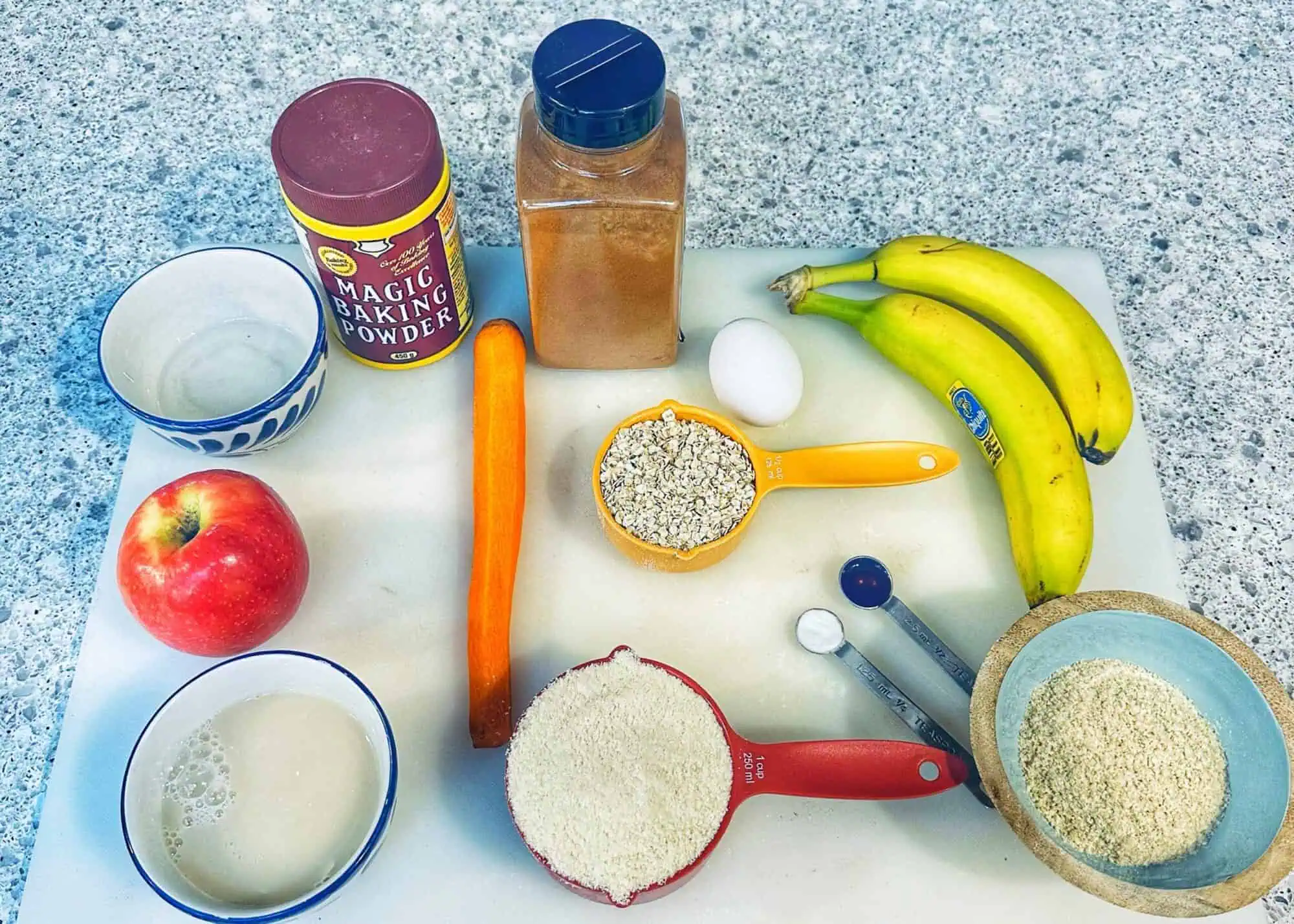 Picture of ingredients for ABC (apple, banana, carrot) muffins for toddlers, placed on a white cutting board on top of the counter.