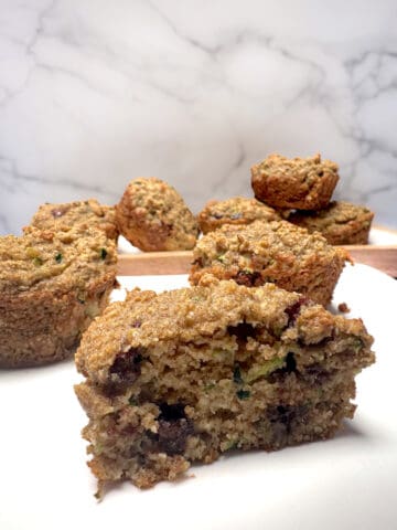 Almond flour zucchini muffins on white plate with front muffin sliced in half and whole muffins in behind.