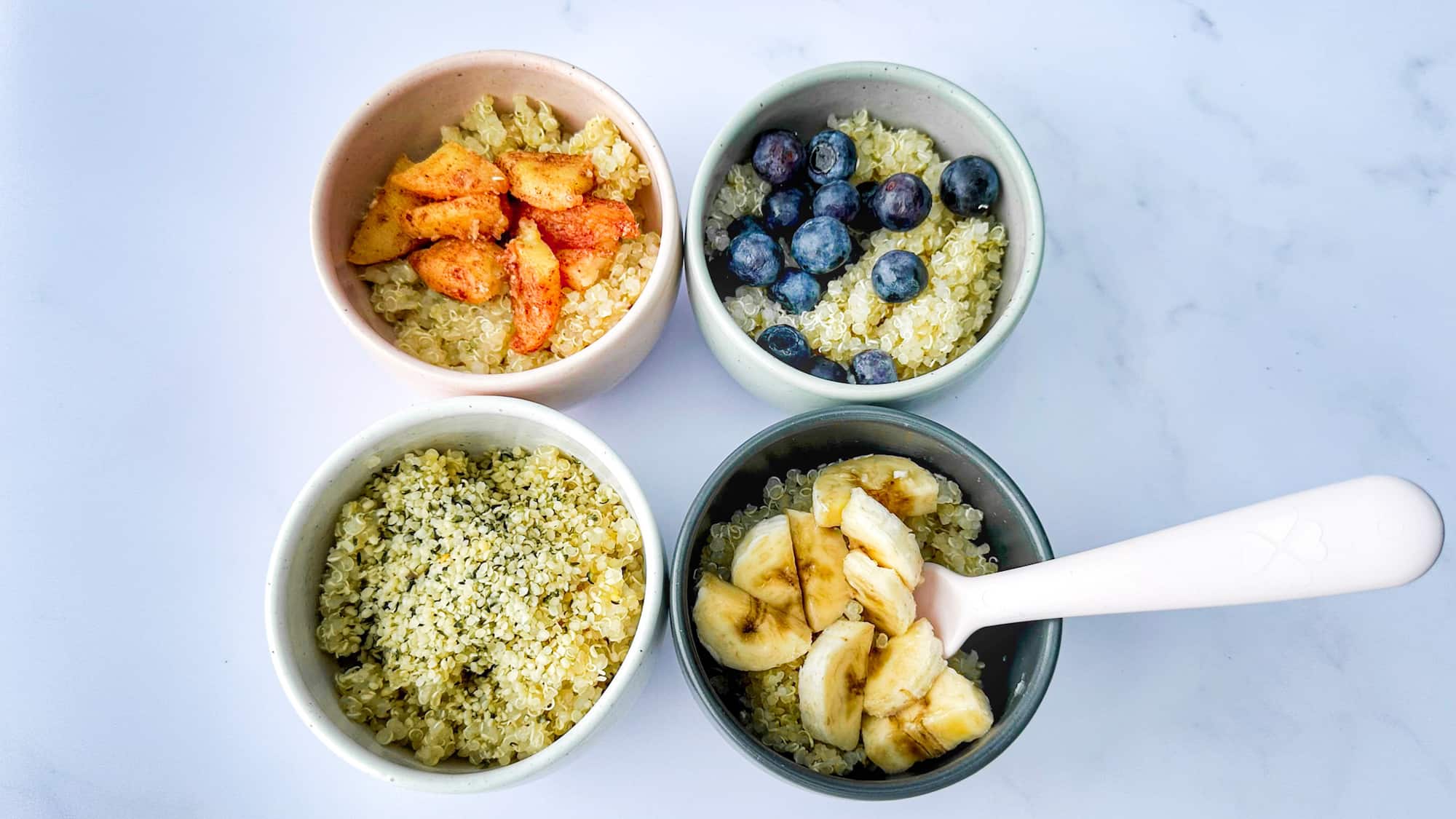 Baby quinoa cereal served in four small bowls with various toppings including sautéed apples, blueberries, hemp hearts and banana with a pink fork.