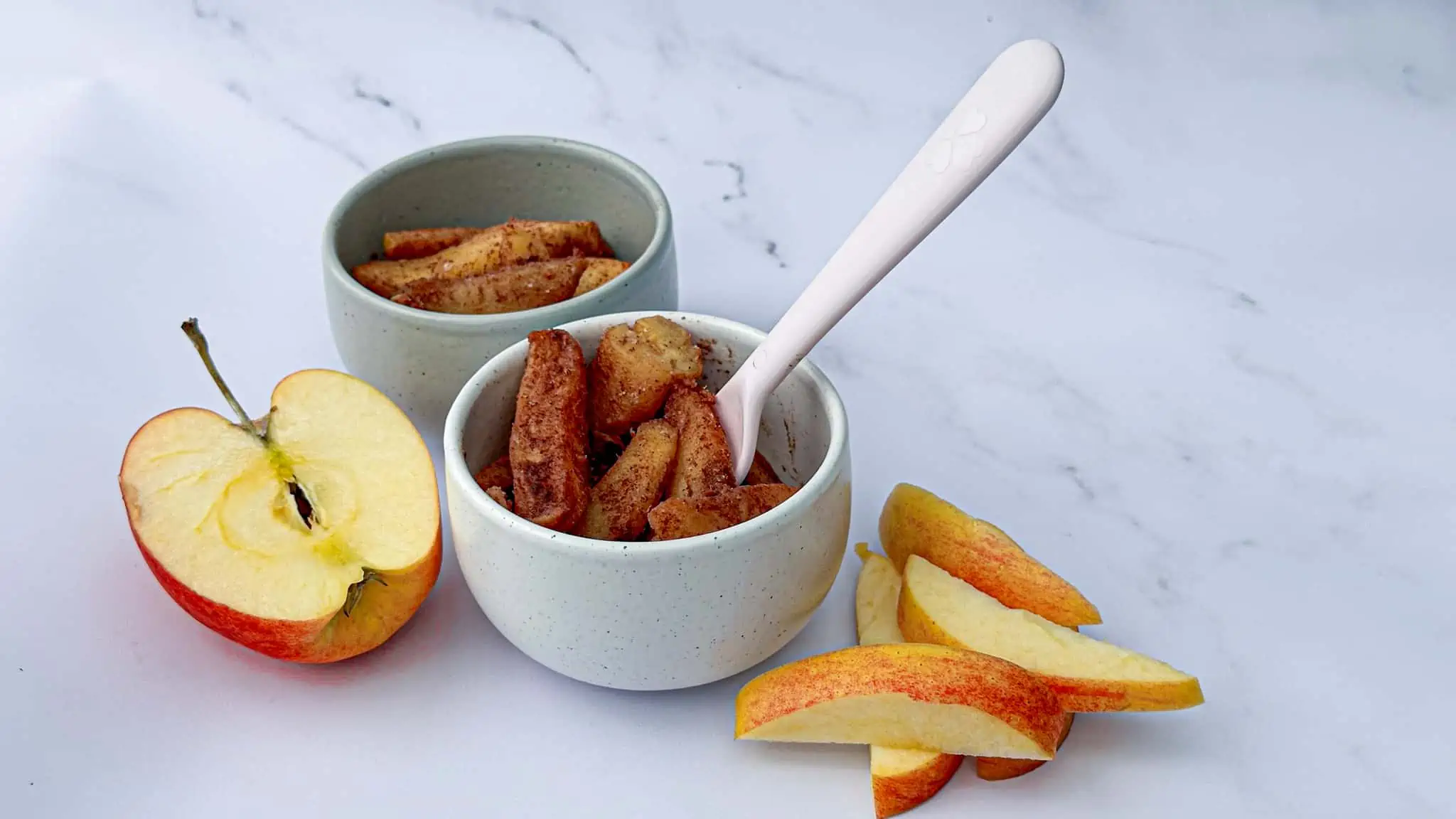 Image of sauteed apples in two white bowls with a pink fork and apple slices on the side.