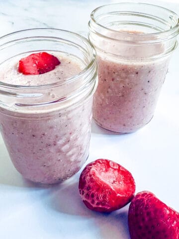 Strawberry smoothie in two glass cups with frozen strawberries on the side.