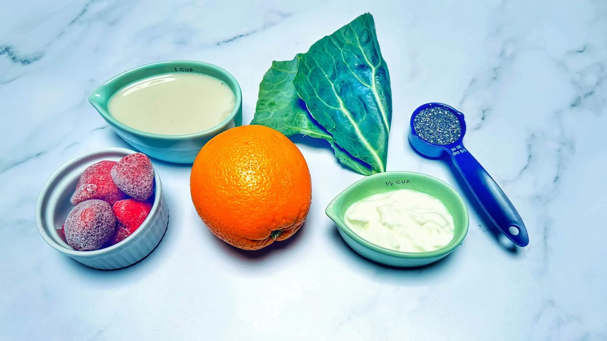 Picture of ingredients for strawberry calcium smoothie including frozen strawberries, almond milk, orange, collard greens, yogurt and chia seeds on marble backdrop.