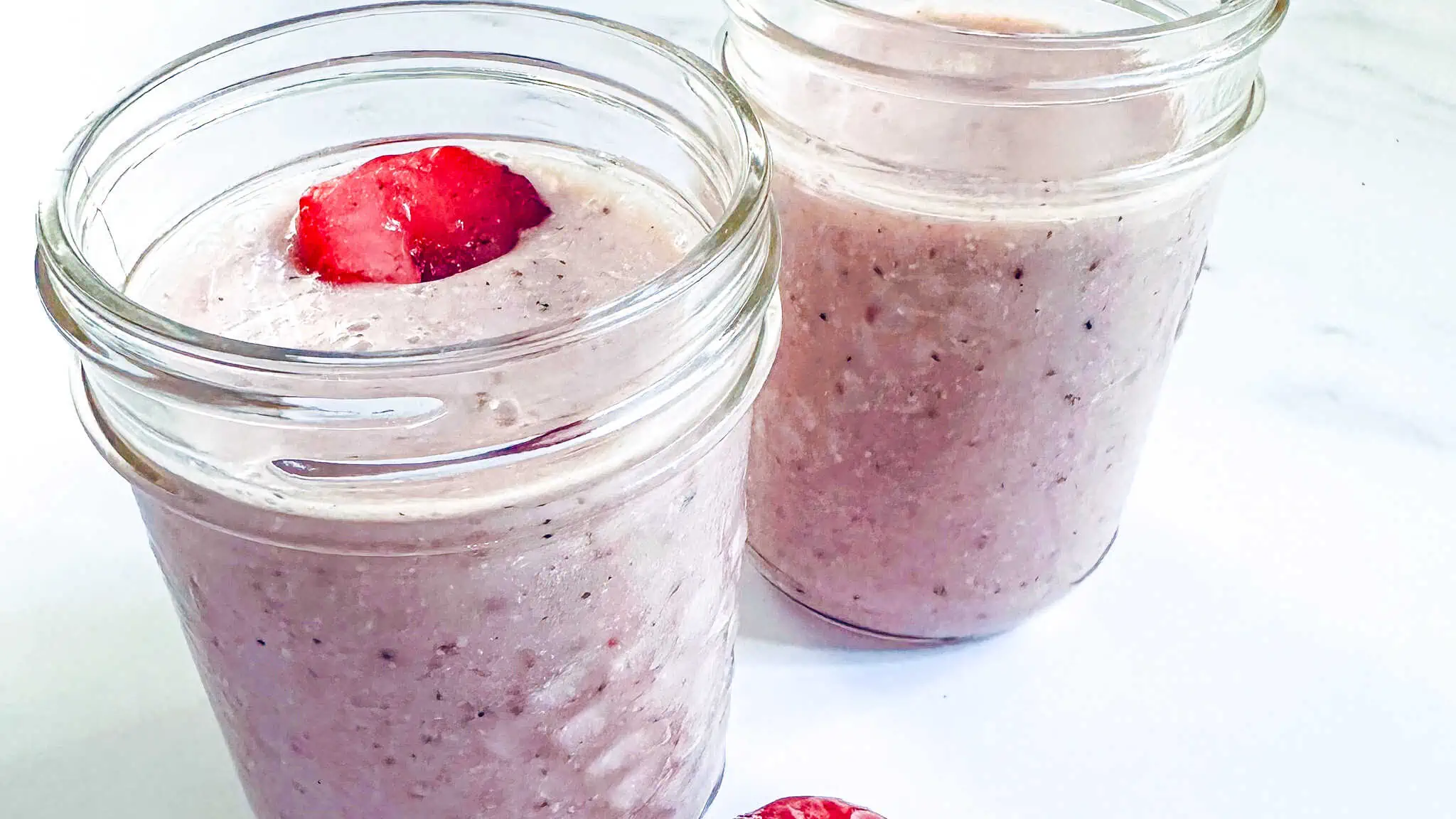 Picture of smoothie in two glass jars on a white marble backdrop with a strawberry in one of the glasses.