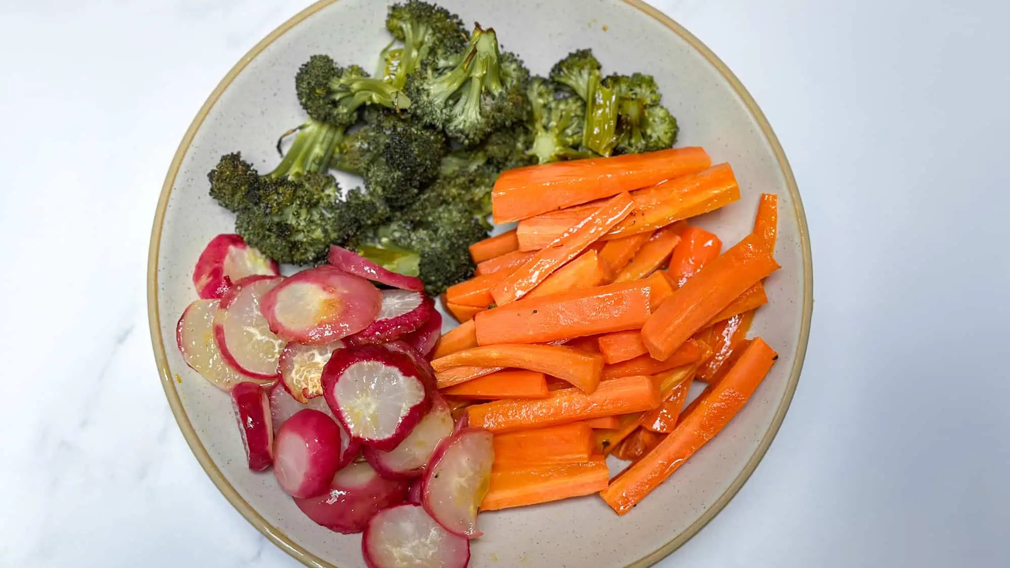 Roasted vegetables including carrots, broccoli and radishes in white bowl on marble background