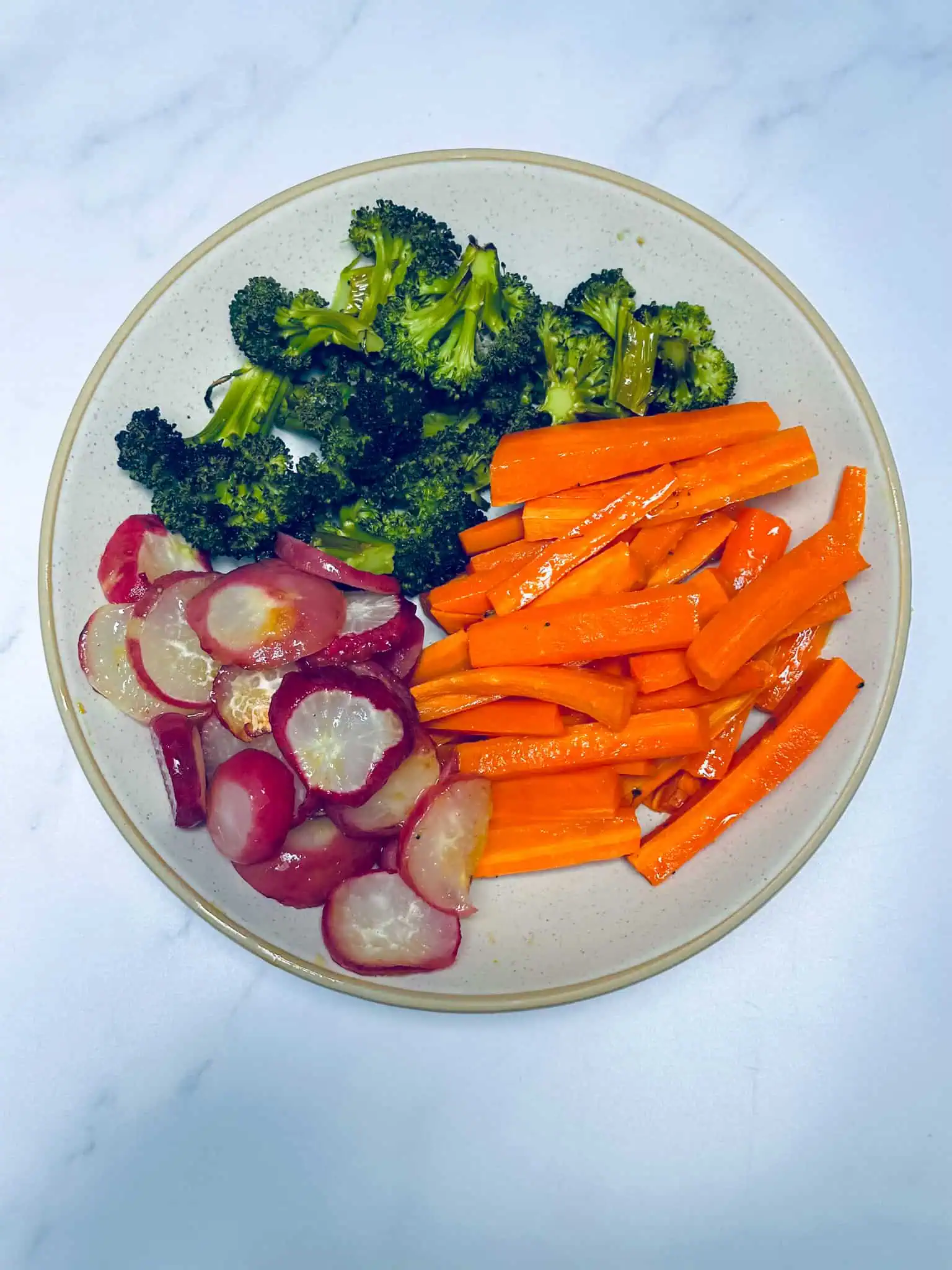 Roasted vegetables including carrots, broccoli and radishes in white bowl on marble backdrop.