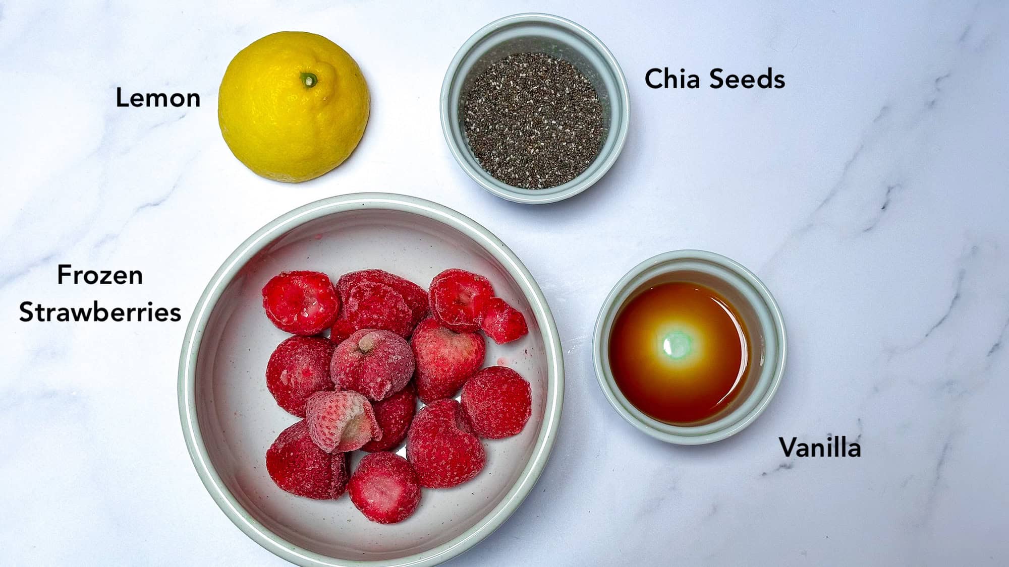 Ingredients for strawberry chia jam including lemon, chia seeds, strawberries and vanilla on white marble backdrop