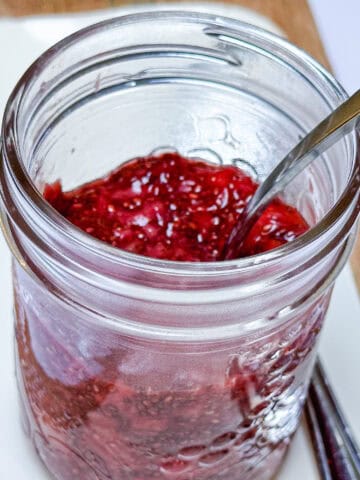 Strawberry chia jam in glass mason jar with silver spoon on white serving plate.