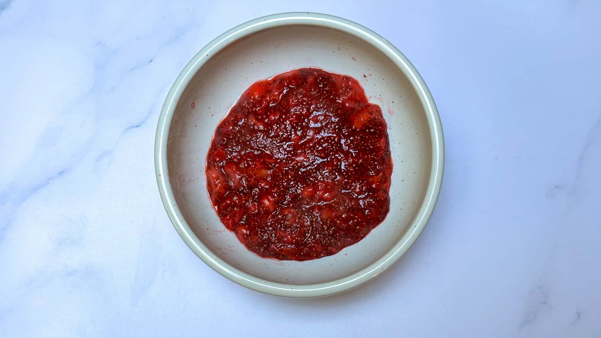 Step 3 of strawberry chia jam, allowing jam to cool in white bowl on white marble backdrop