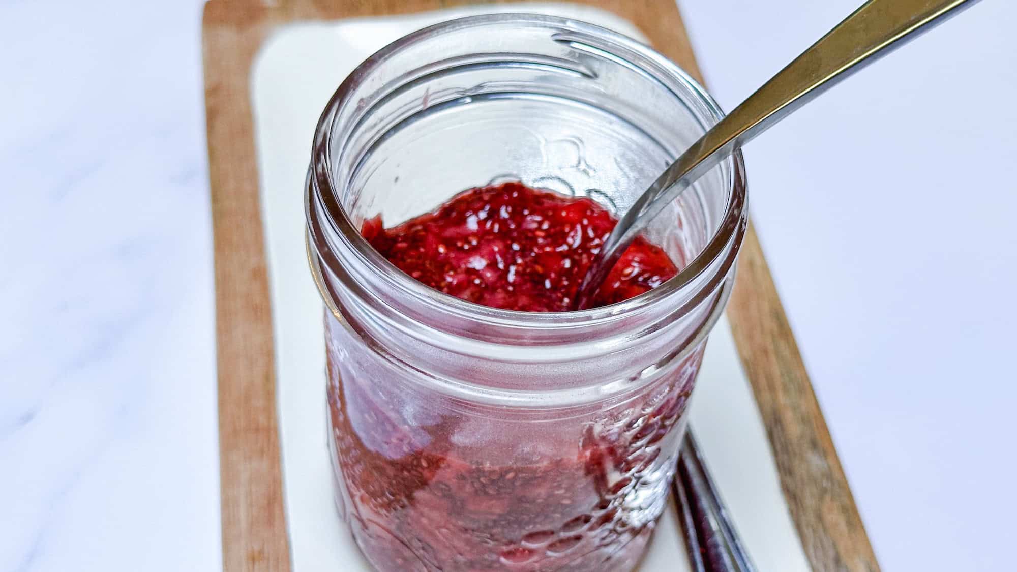 Strawberry chia jam in glass mason jar with silver spoon on white and wood serving plate.