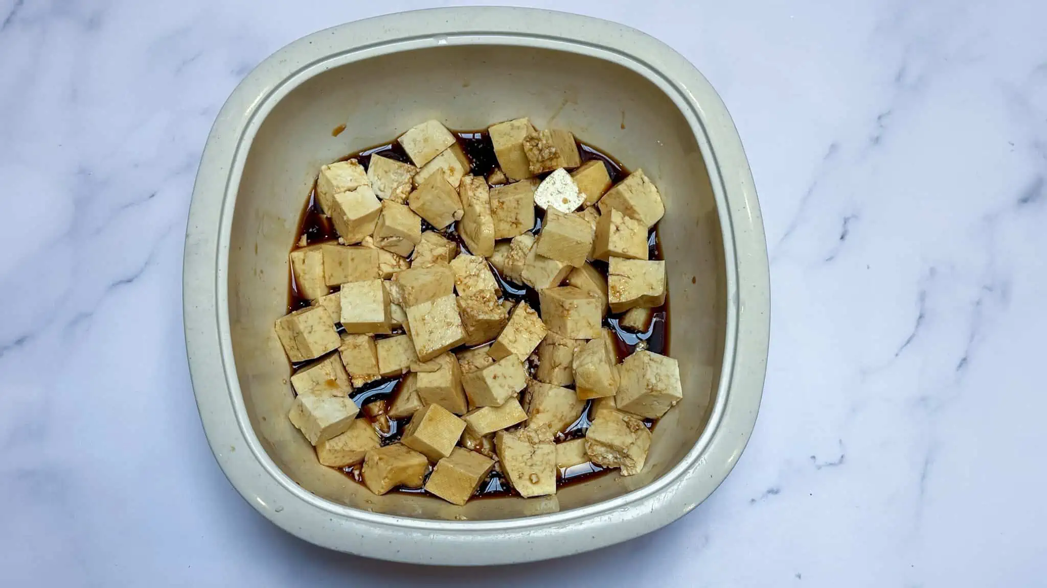 Step 2 of tofu rice bowls, marinading tofu in white plastic bowl on white marble backdrop.