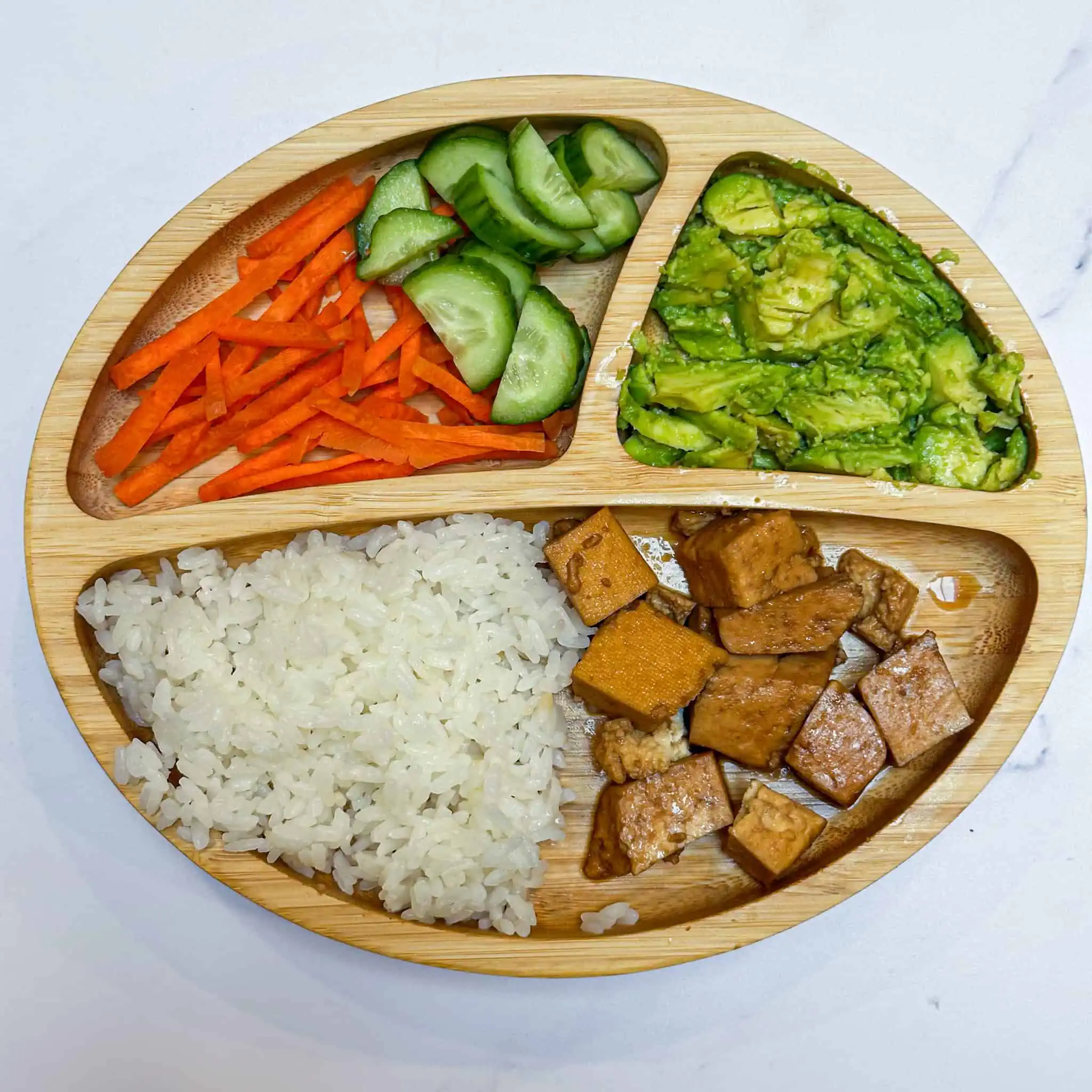 Image of tofu rice bowl deconstructed onto wooden toddler plate on white marble backdrop.