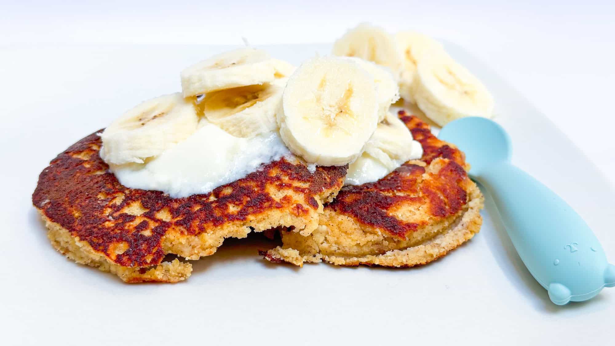 Image of almond flour oat pancakes on white plate topped with yogurt and banana and blue baby spoon next to pancake