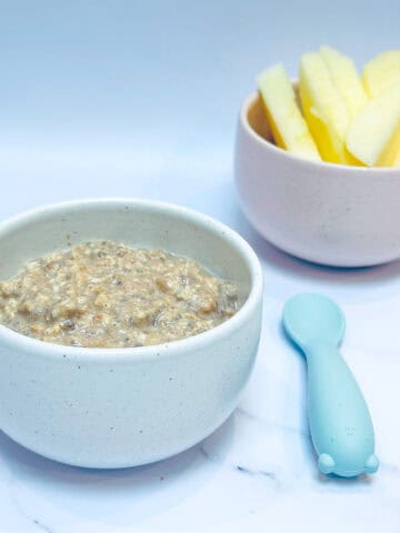 Applesauce overnight oats in small white bowl with blue baby spoon and bowl of apple slices in background.