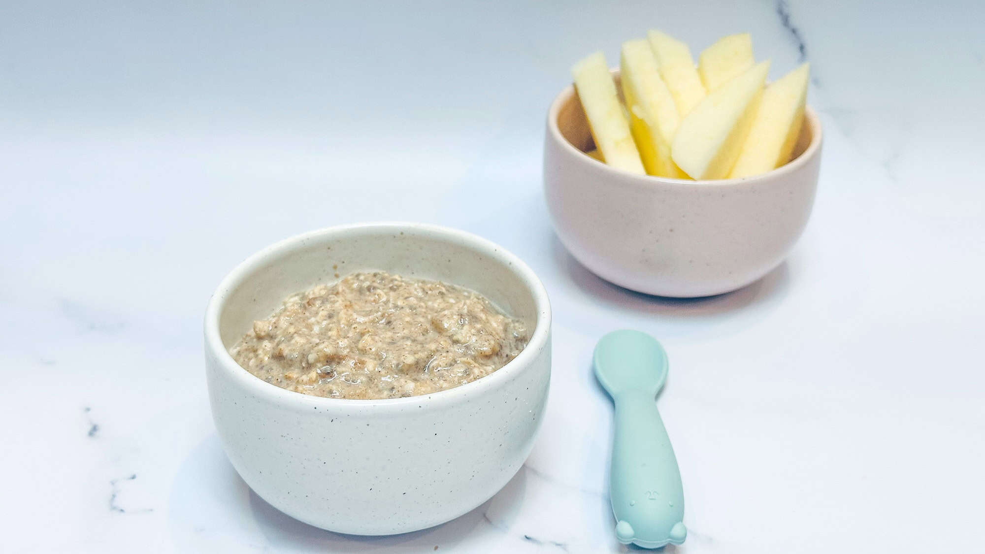 Applesauce overnight oats in white bowl with blue baby spoon and cut up apples in background.