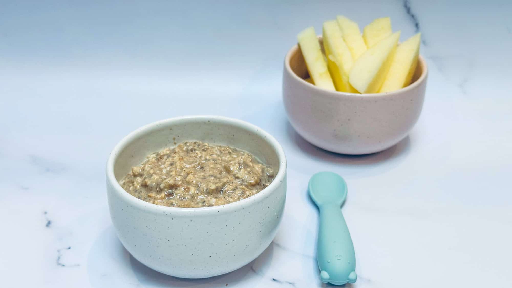 Applesauce overnight oats in small white bowl with blue baby spoon and bowl of apple slices in background.