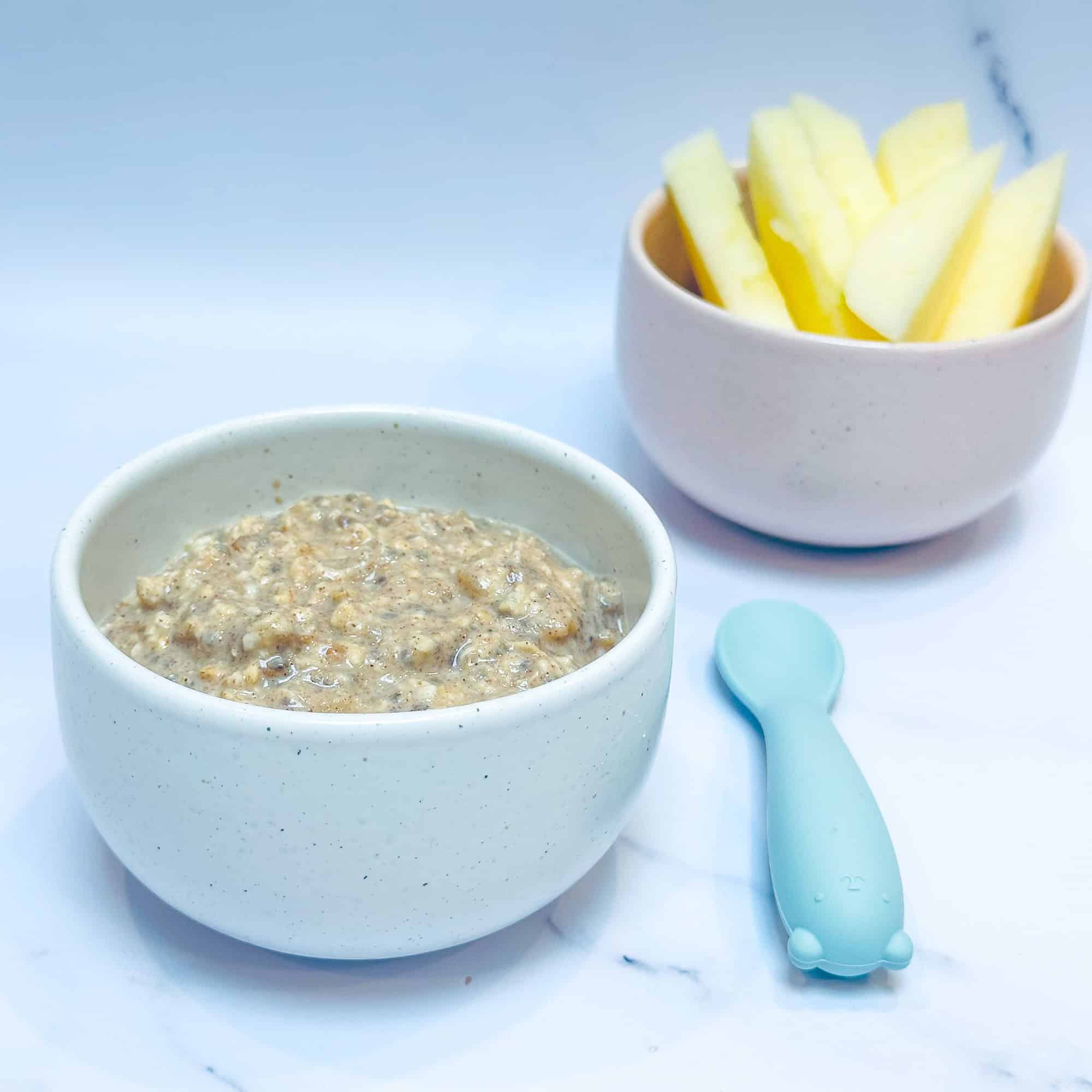 Applesauce overnight oats in small white bowl with blue baby spoon and bowl of apple slices in background.