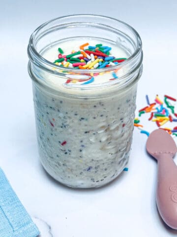 Square image of birthday cake overnight oats in glass mason jar with sprinkles on top and pink baby spoon to the side.