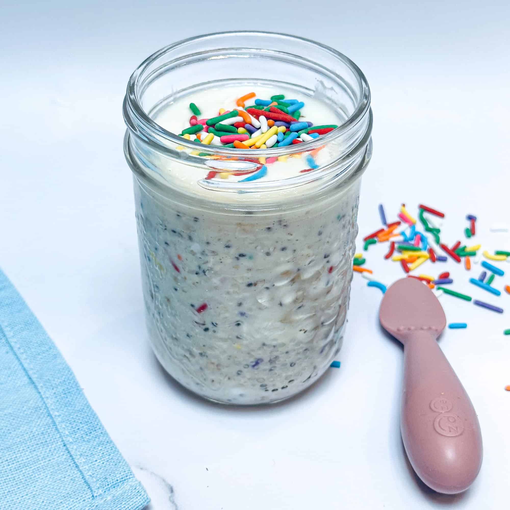 Square image of birthday cake overnight oats in glass mason jar with sprinkles on top and pink baby spoon to the side.