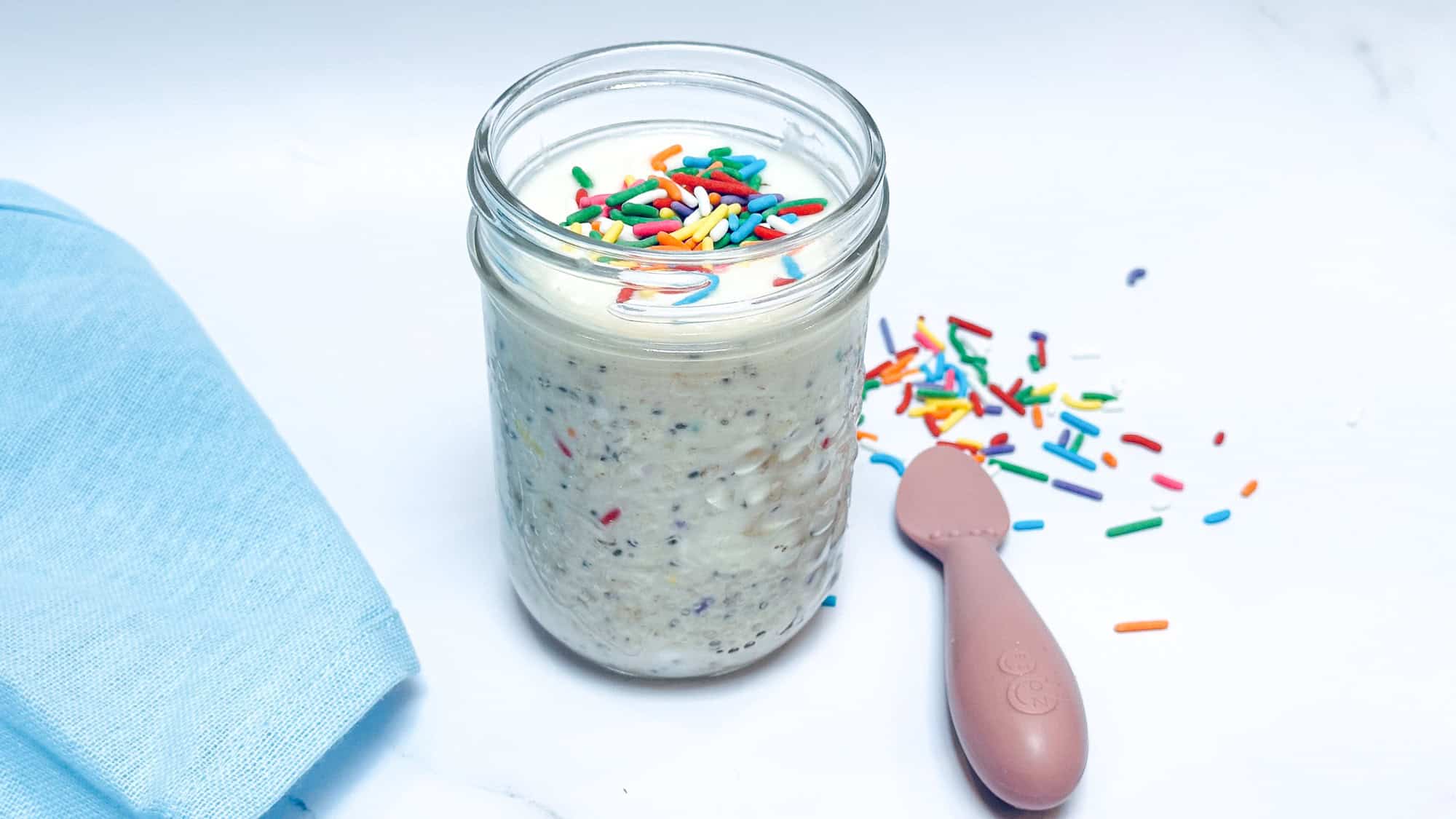 Image of Birthday Cake Overnight Oats in glass mason jar with sprinkles on top and pink baby spoon to the side.