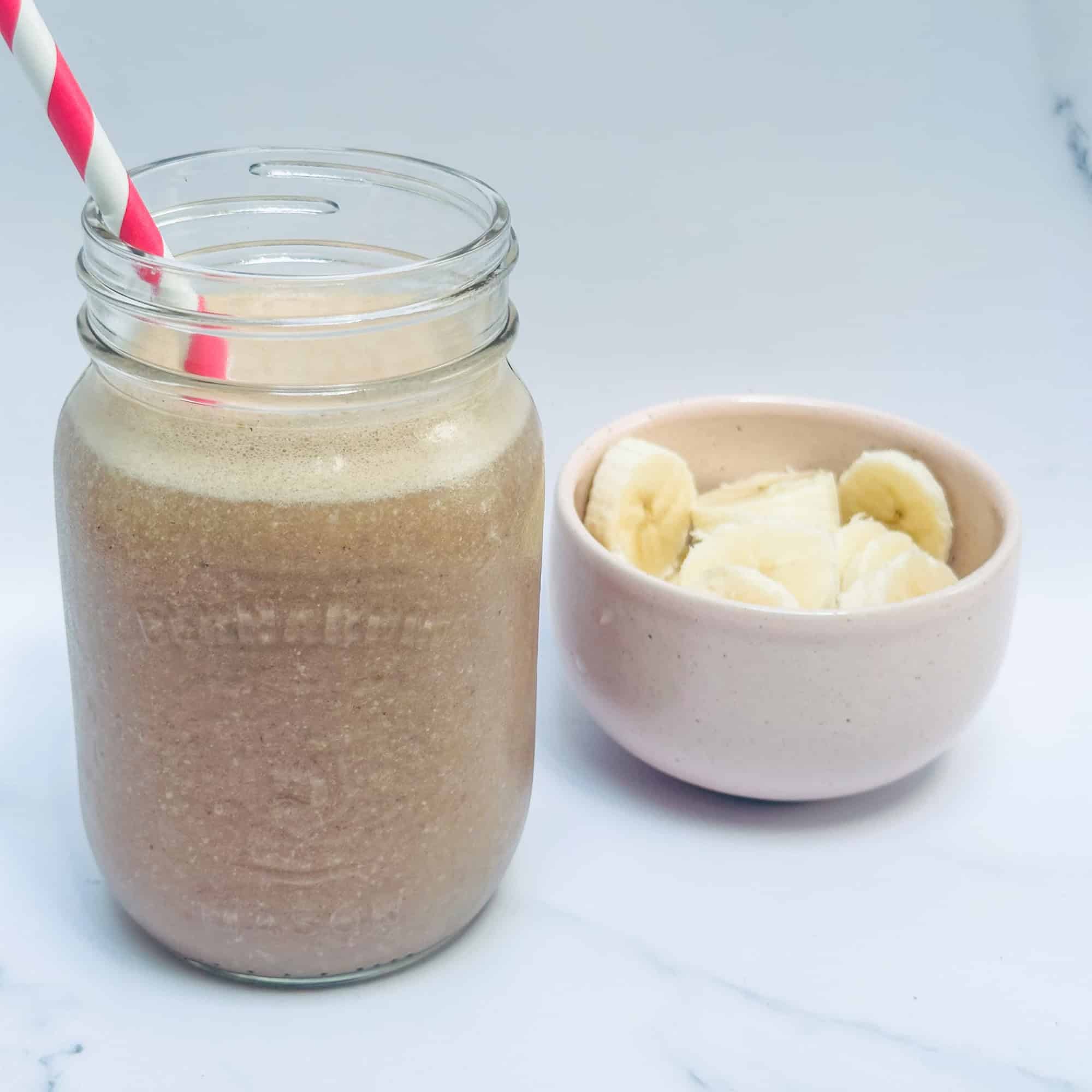 Chunky Monkey smoothie in glass mason jar with pink straw and cut up bananas in background in small pink bowl.