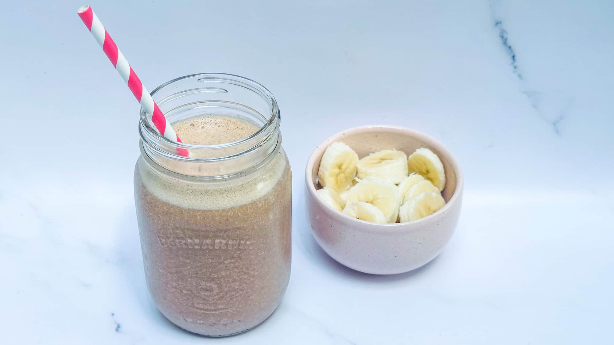 Chunky Monkey Smoothie in glass jar with pink striped straw and bowl of banana slices in background.