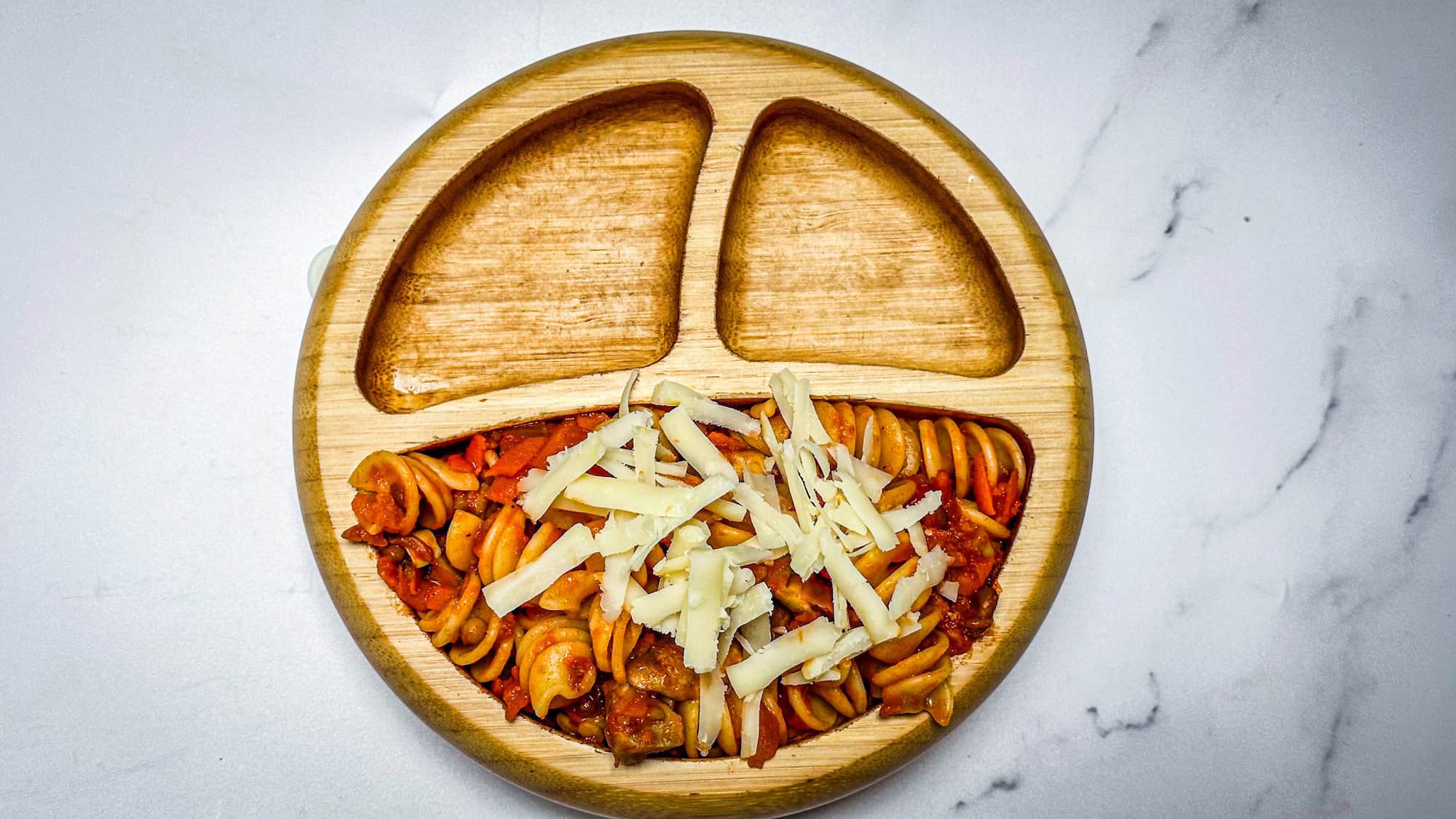 Pasta with lentil sauce in wooden toddler bowl on white marble background.