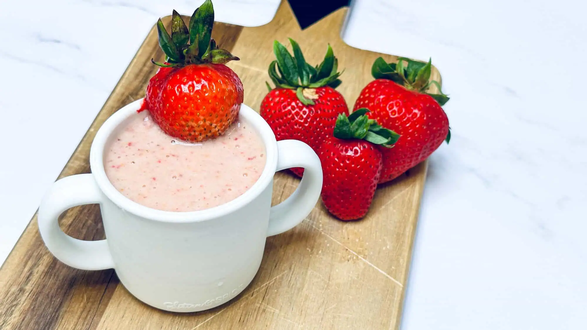 Strawberry peanut butter smoothie in white silicone toddler cup with handles on wooden cutting board.