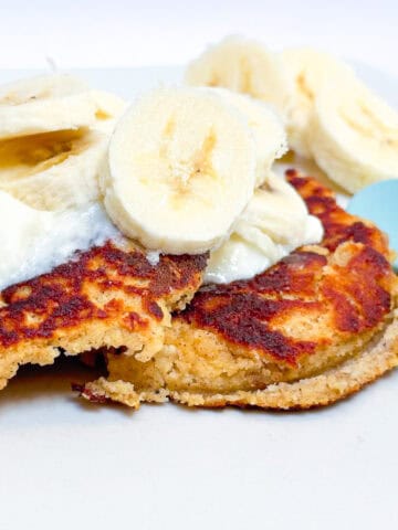 Image of almond flour and oatmeal pancakes topped with yogurt and banana on a white plate with a blue toddler spoon