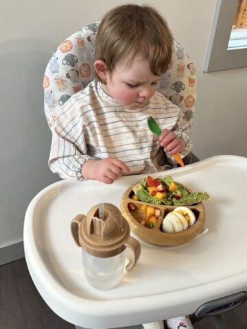 Image of child sitting in high chair eating out of wooden bowl with a fork.