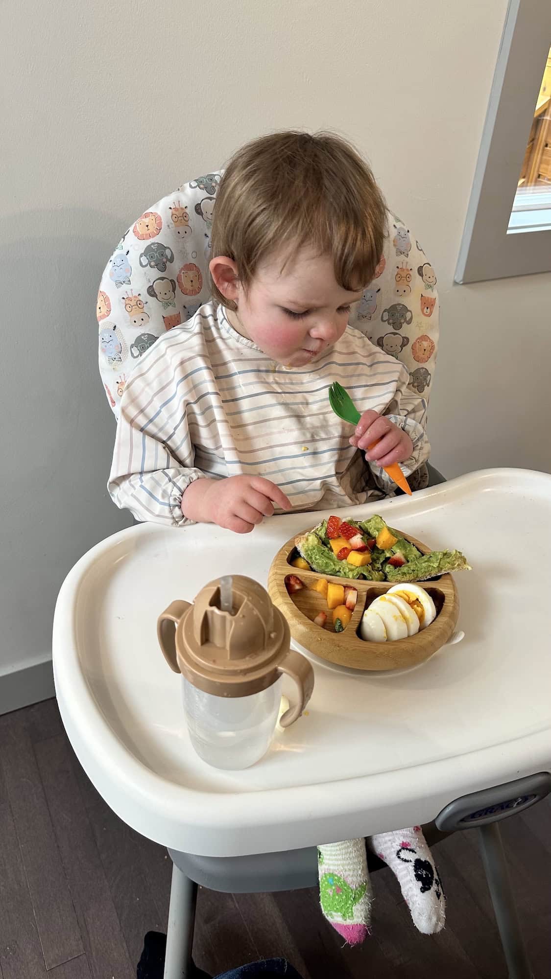 Image of child sitting in high chair eating out of wooden bowl with a fork.