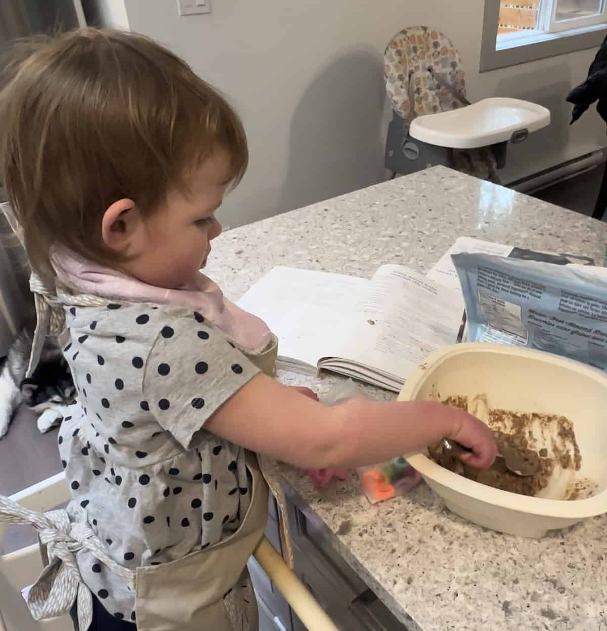 Image of child standing in toddler tower stirring cookie dough in white bowl.