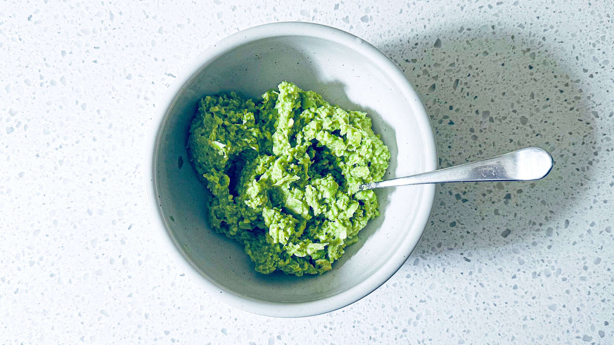 High protein guacamole in a white bowl with a silver spoon on white marble background.