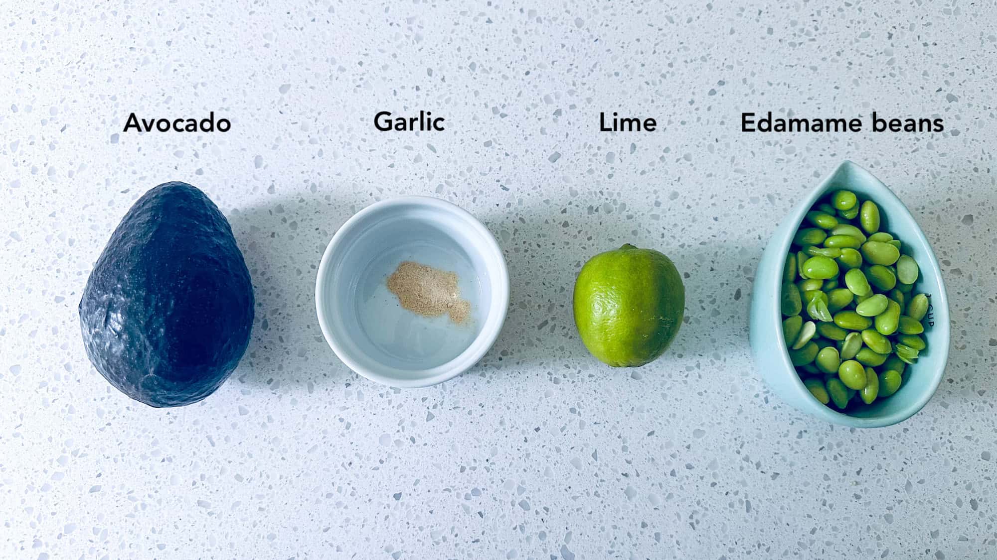 Ingredients for high protein guacamole. Avocado, garlic, lime and edamame beans in small bowls on white marble background.