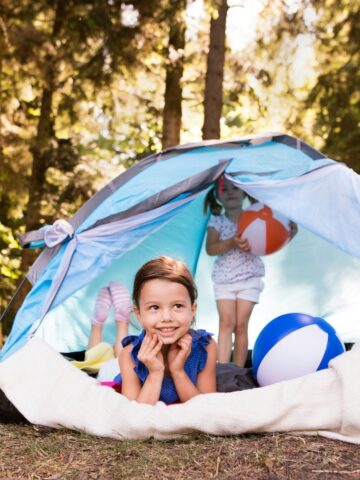 Image of two kids in a tent camping. One child is holding an orange and white ball and the other child is lying down looking out the tent.