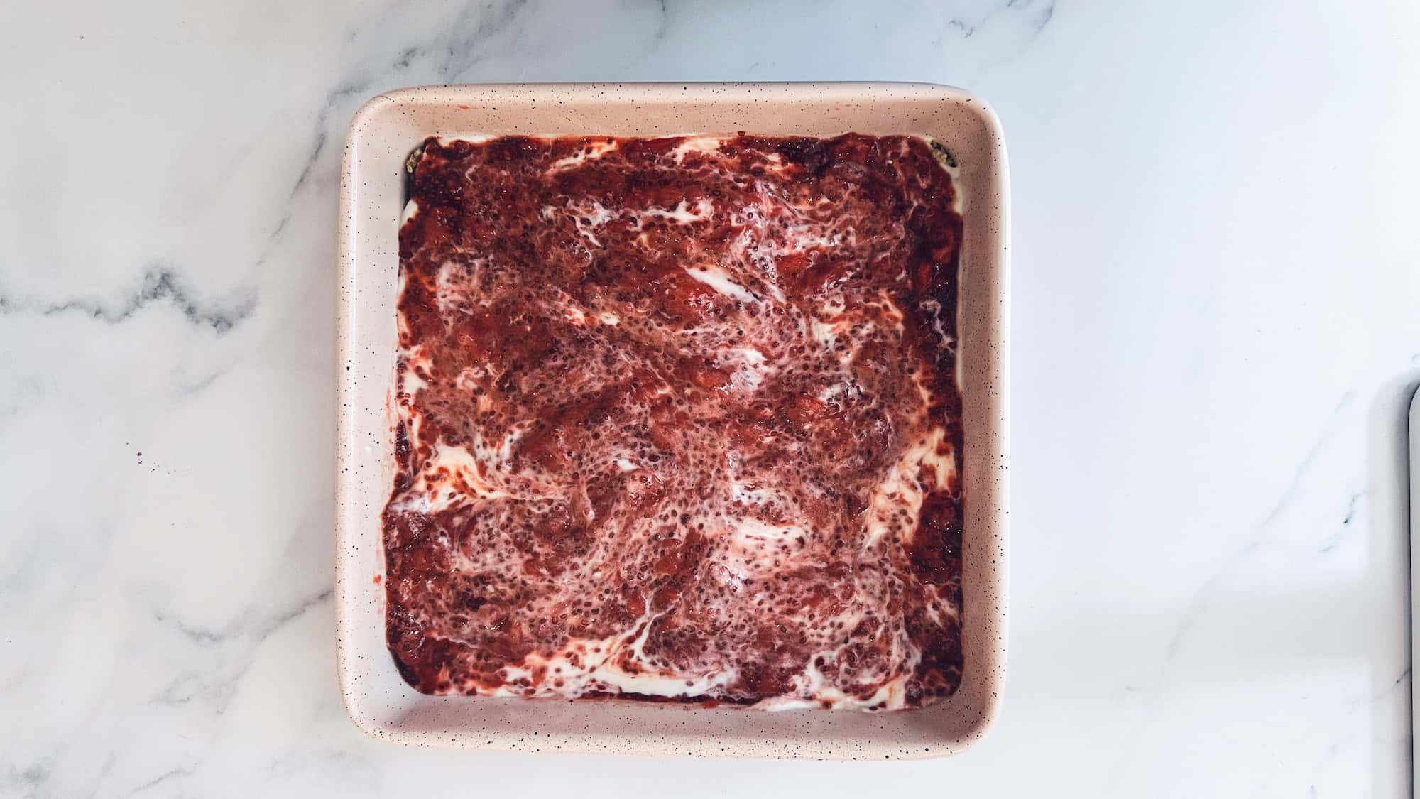 Step 4 for strawberry yogurt protein bars. Yogurt and strawberry chia jam are added on top of crust in a square pink pan on a white marble background.