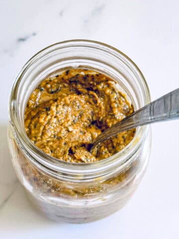 Nut and seed butter in glass mason jar with silver spoon on white marble backdrop.