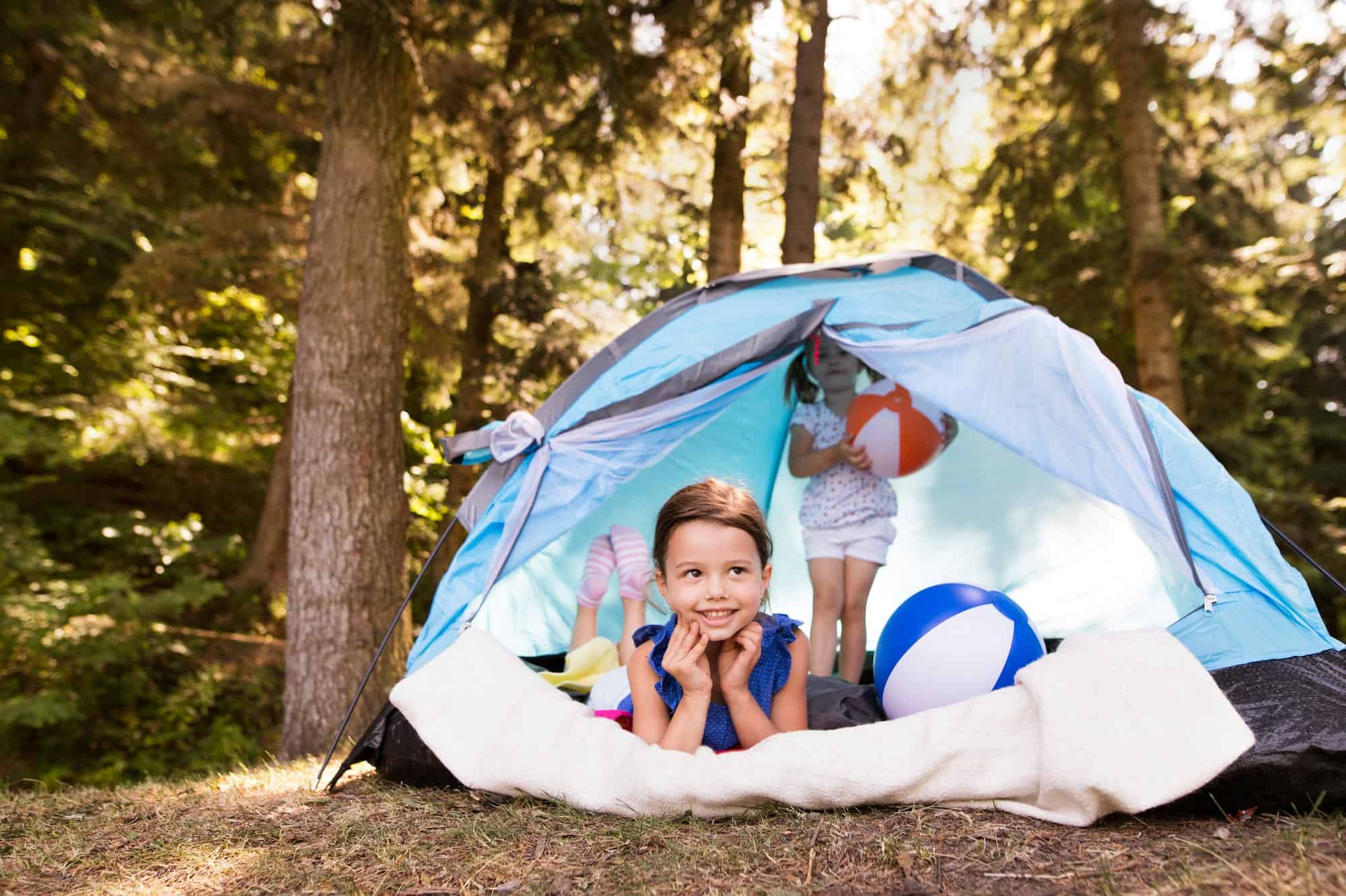 Image of two young children in a tent in the forest. One child is holding a orange and white ball and the other child is lying down looking out of the tent.