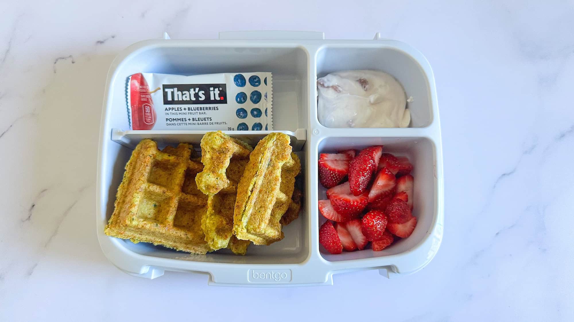 Packed lunchbox in bento box style with savoury waffles, strawberries, yogurt and fruit bar