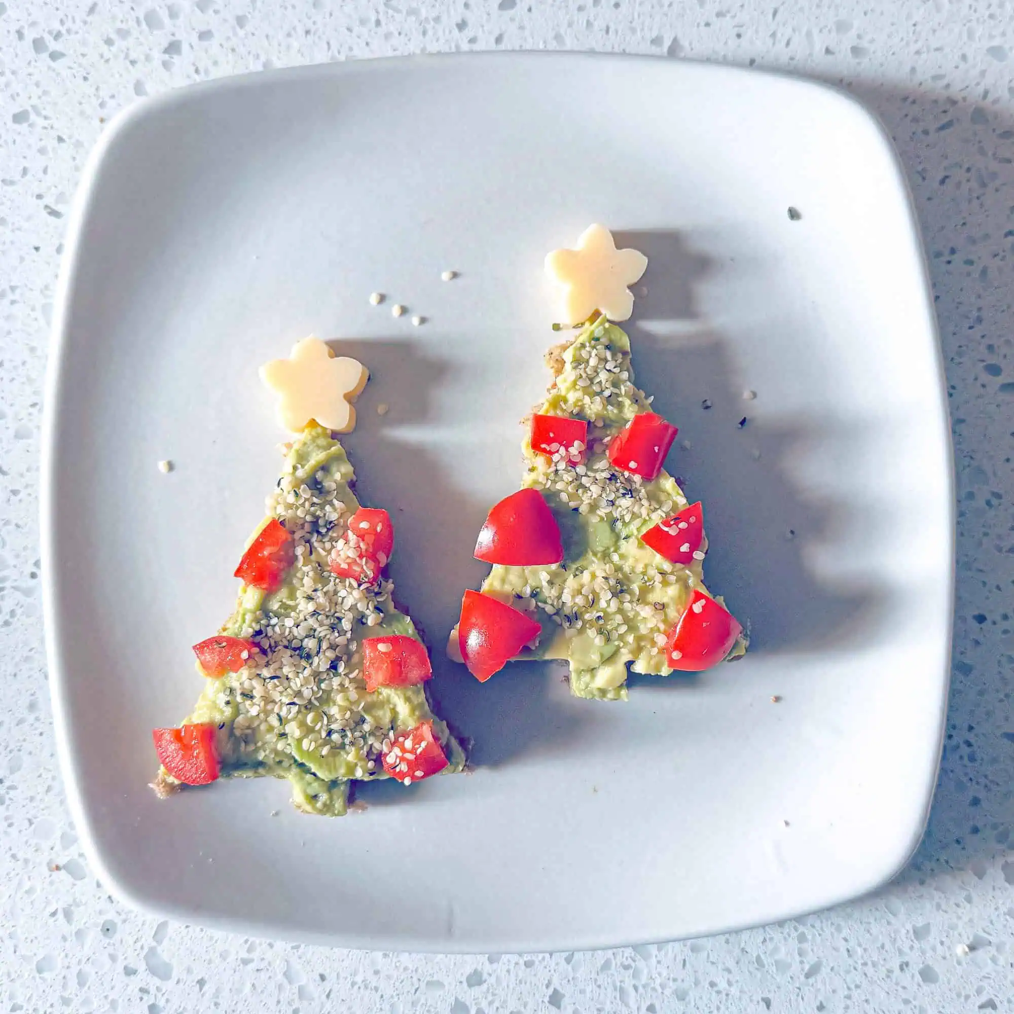 Two Avocado toast Christmas trees on white plate, decorated with tomatoes, red peppers and cheese star.