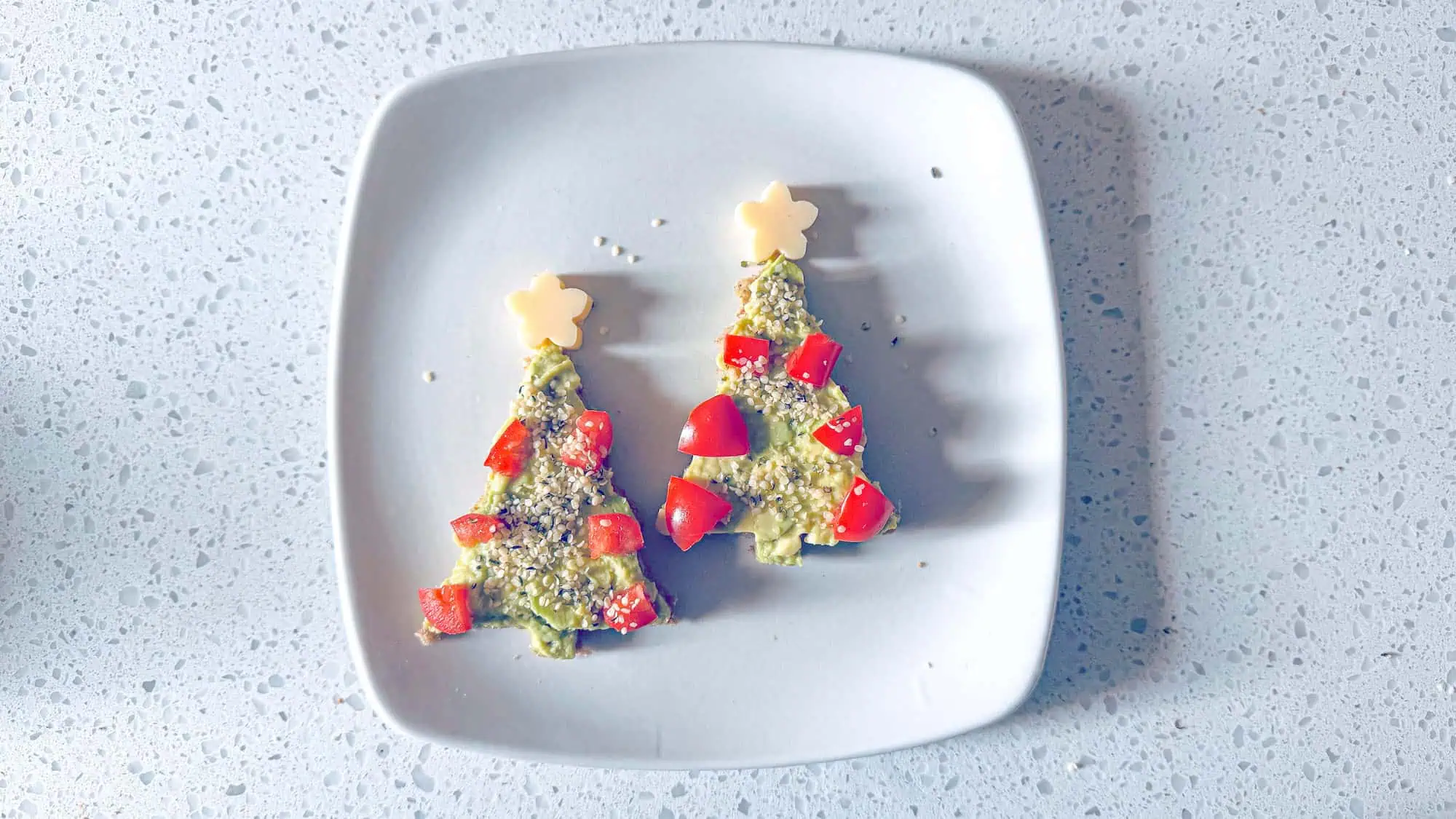 Two avocado toast Christmas Trees on white plate decorated with red tomatoes and cheese star.