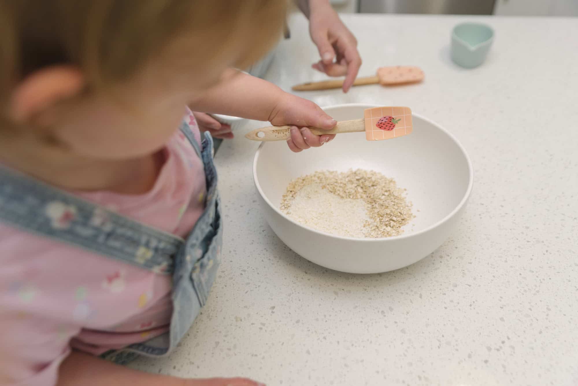 Image of child holding mixing spoon over a white bowl with oats and almond flour in bowl at kitchen counter.