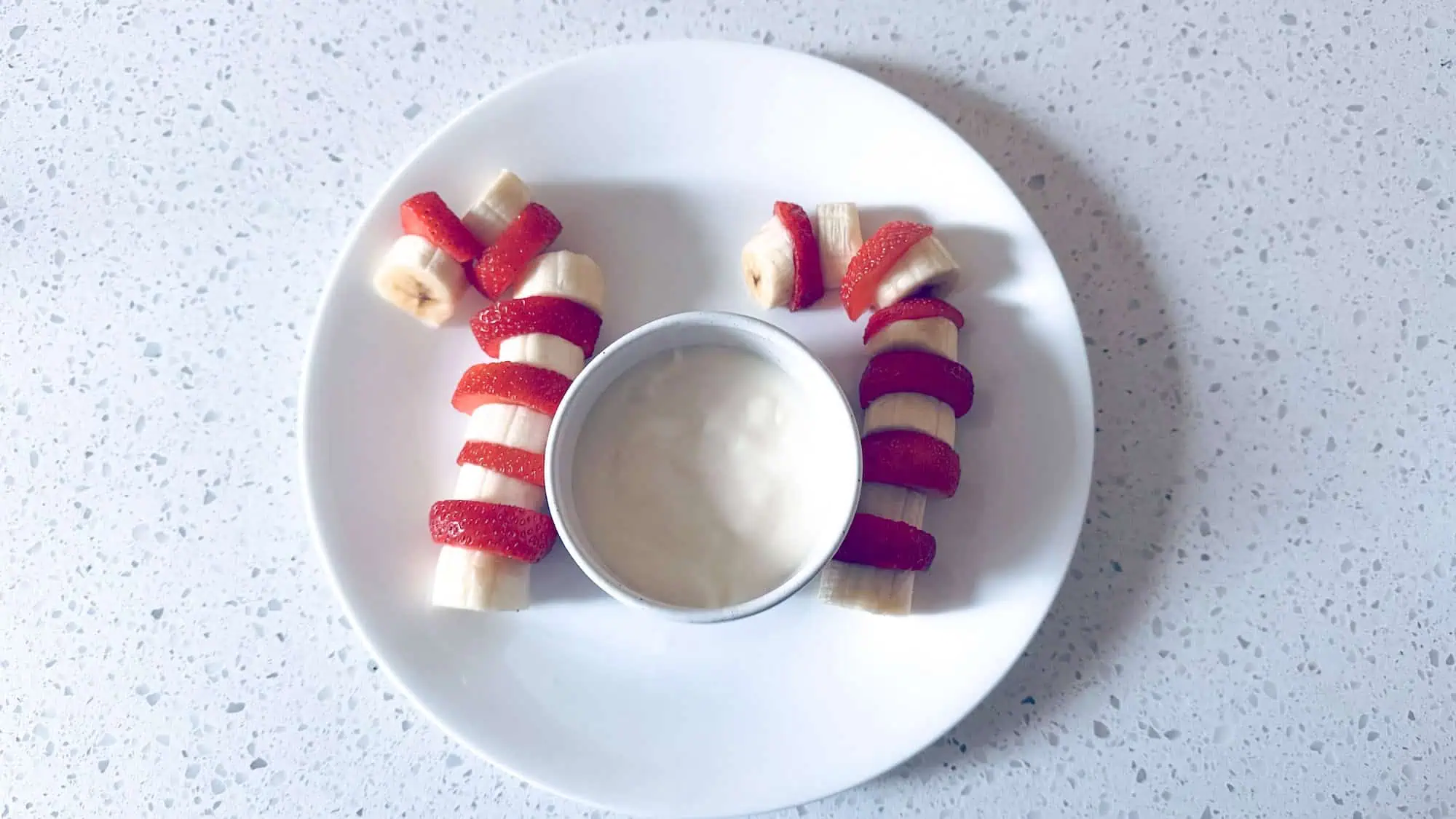 Fruit candy canes made from strawberry and banana with yogurt dip in the middle in a white bowl.