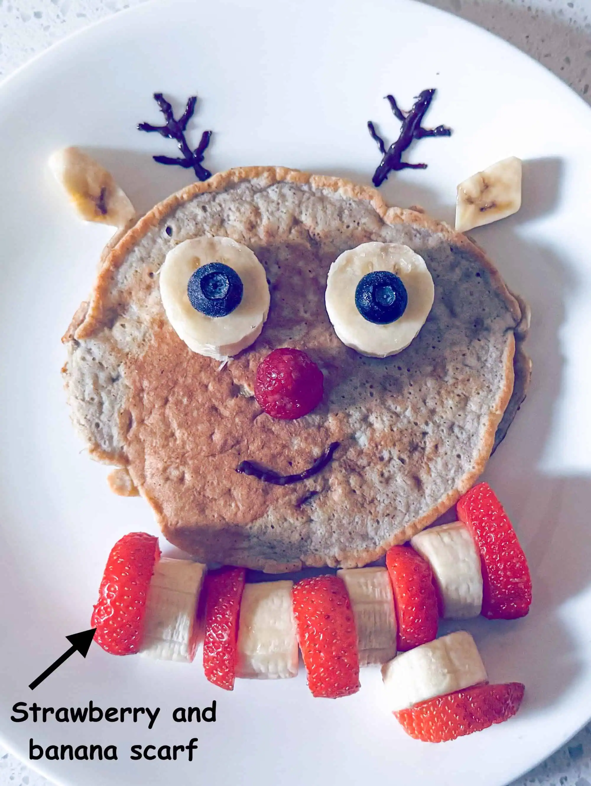 Reindeer pancake with strawberry and banana scarf on white plate.