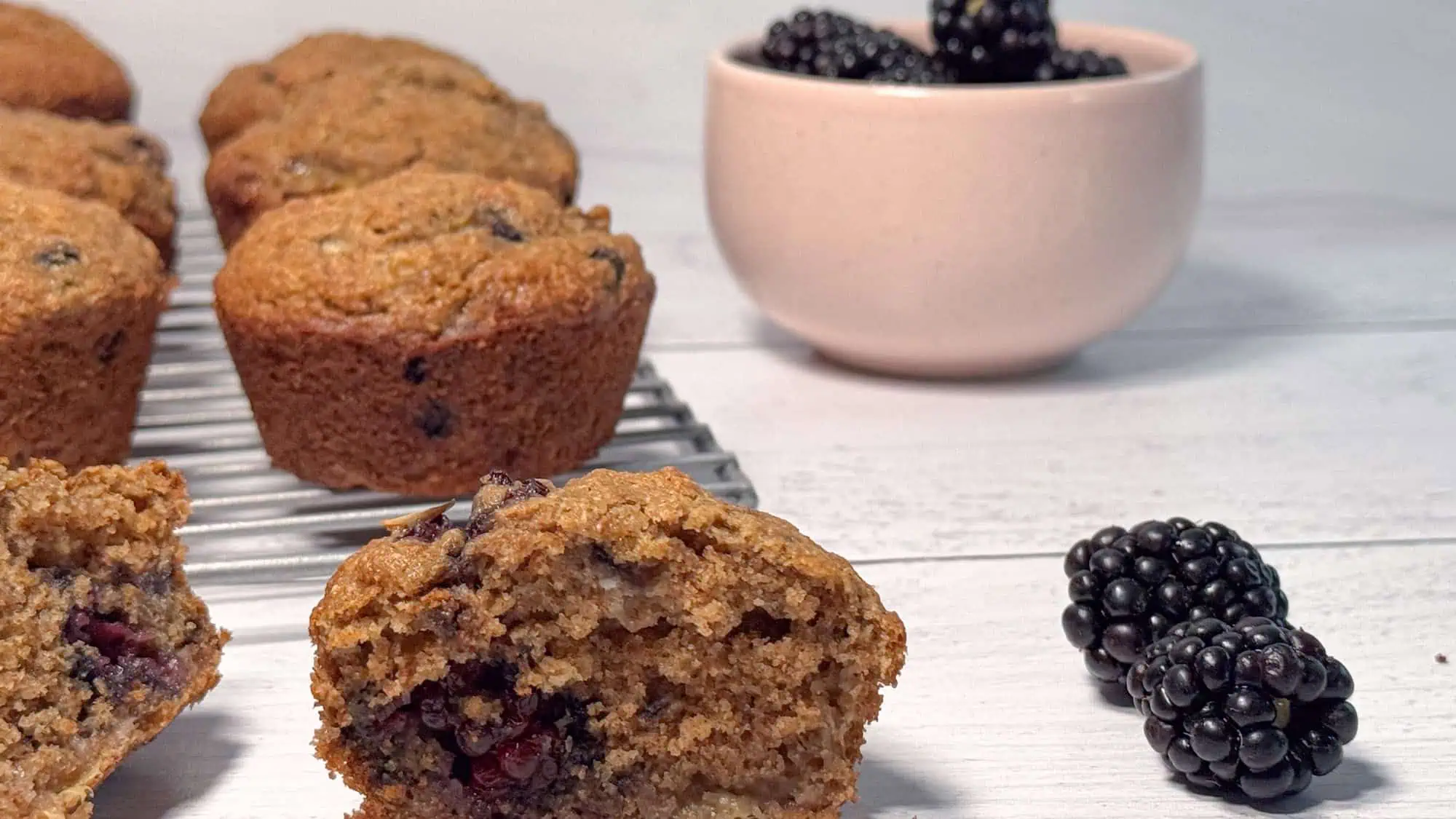 Banana blackberry muffins on white shiplap, with muffins on wire cooling tray and blackberries in pink bowl in background.