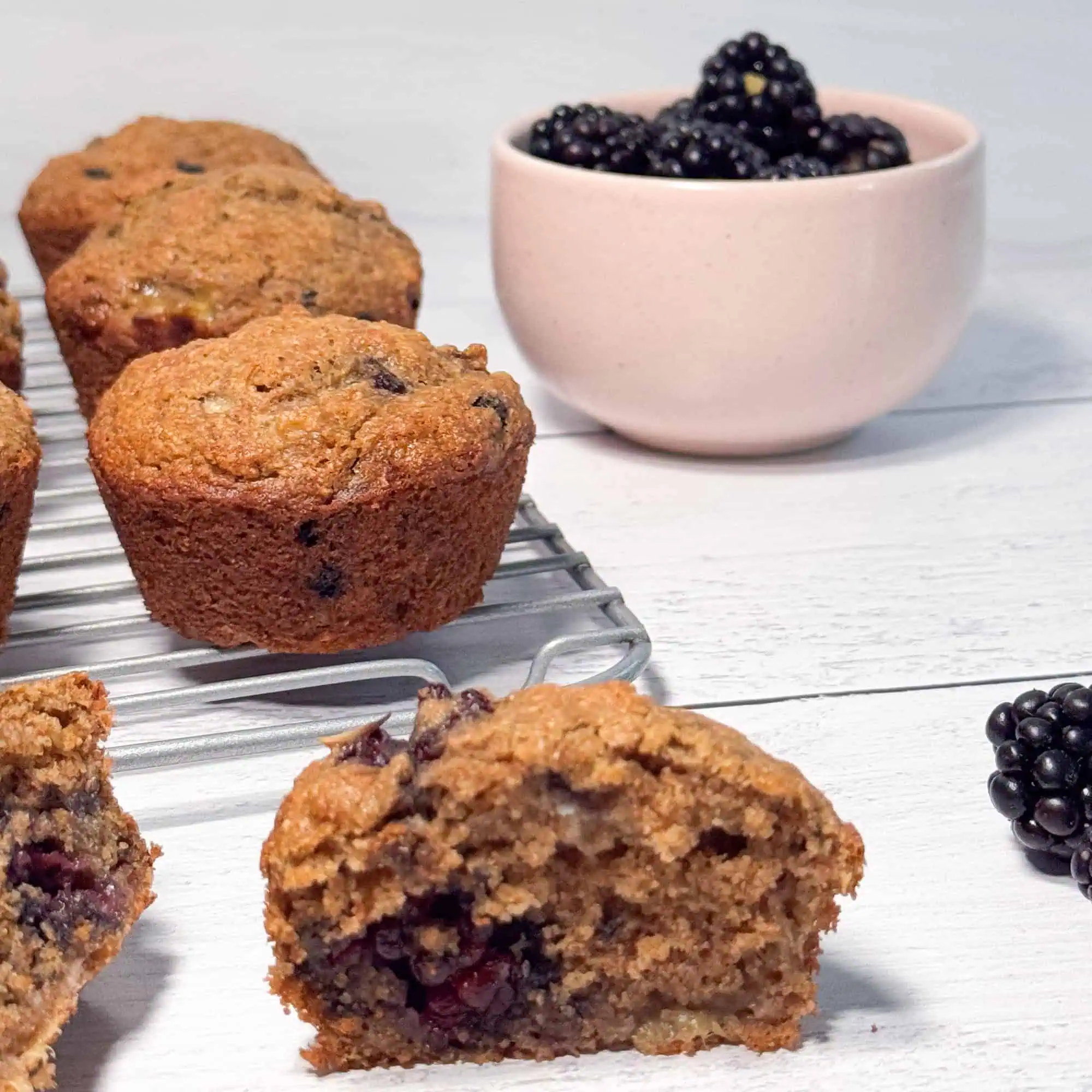 Banana blackberry muffins on white shiplap with blackberries in a pink bowl.