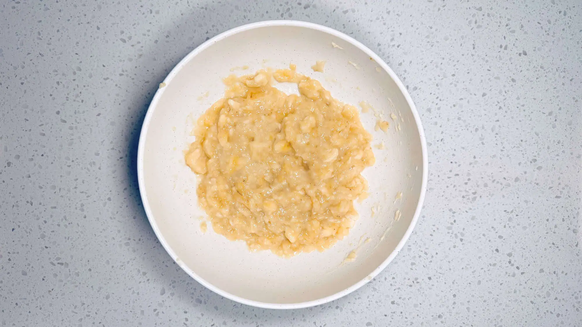 Step 2 of muffins, bananas are mashed and wet ingredients are mixed together in a separate white ceramic bowl.