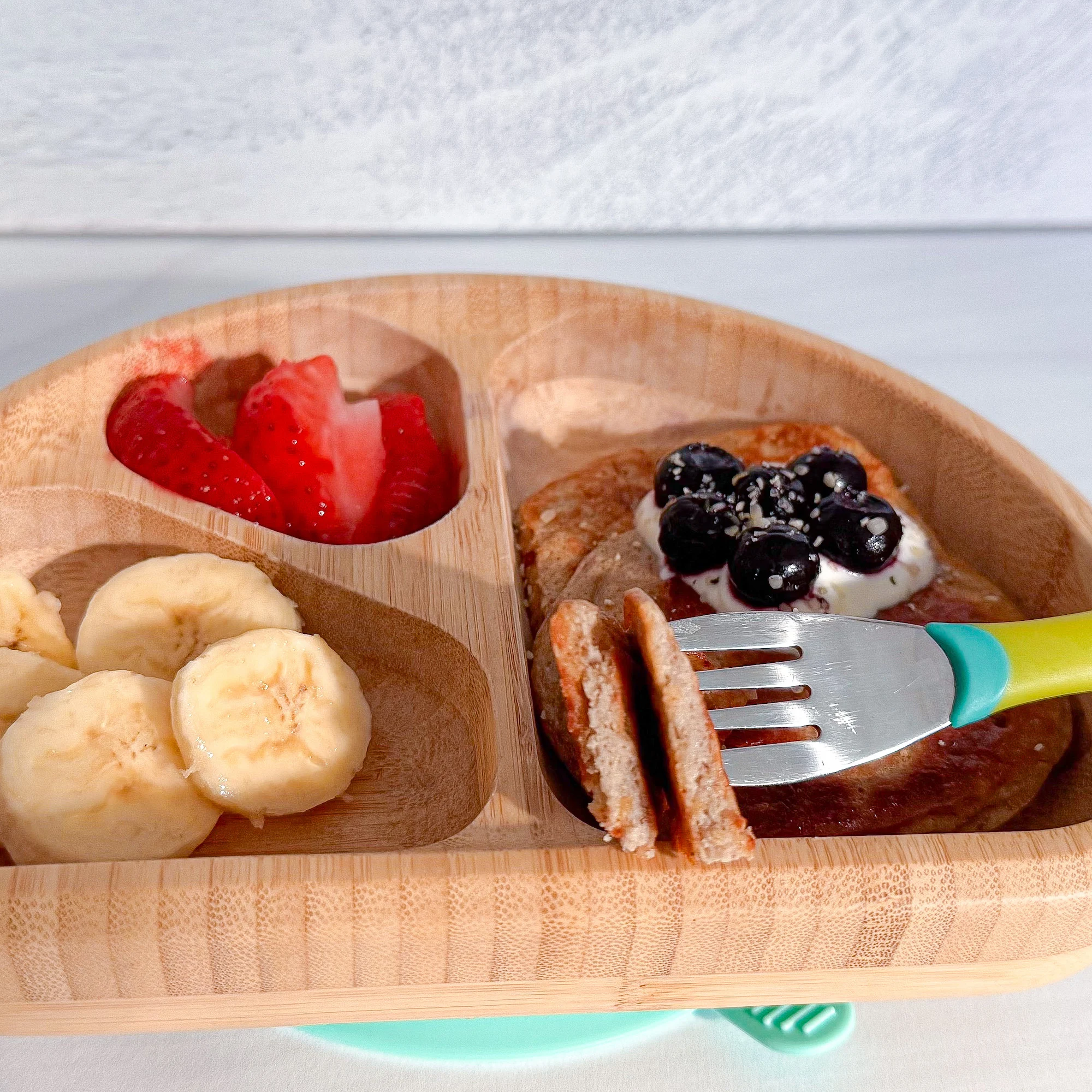 Cottage cheese pancakes in wooden toddler bowl topped with blueberries and side of banana and strawberries