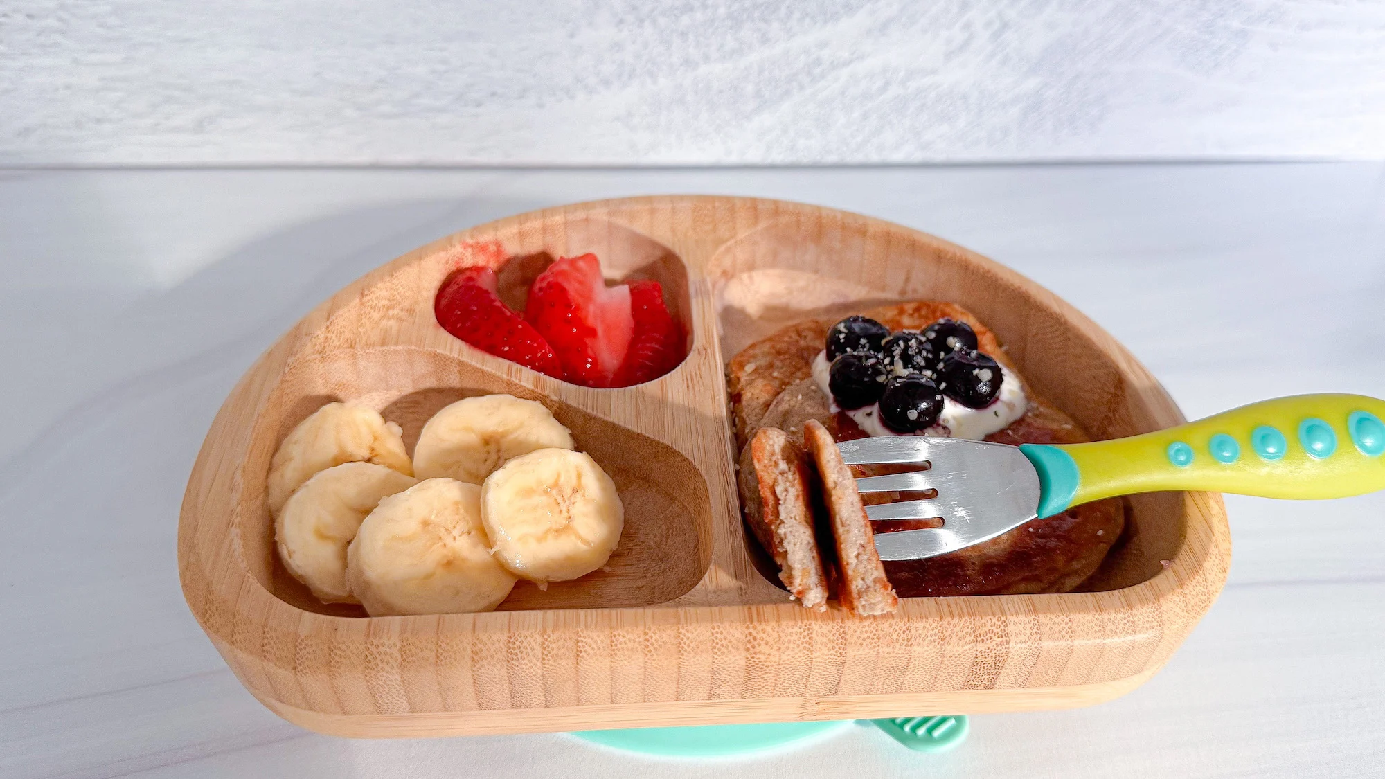 Cottage cheese banana pancakes in wooden toddler bowl topped with blueberries and side of strawberries and banana.