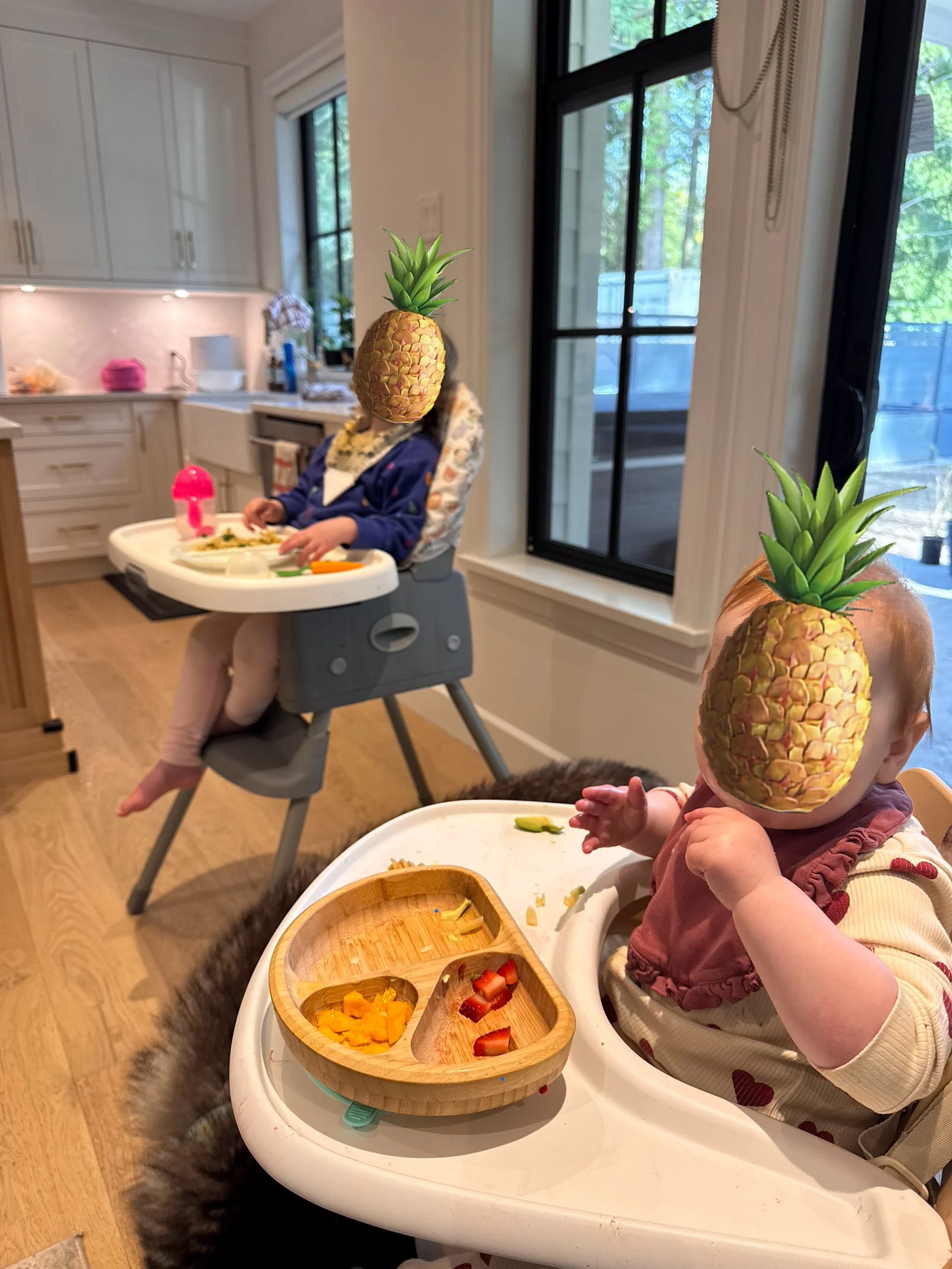 10 month baby sitting in high chair eating dinner with her toddler sister in her high chair eating her dinner. Both faces are covered with pineapples.