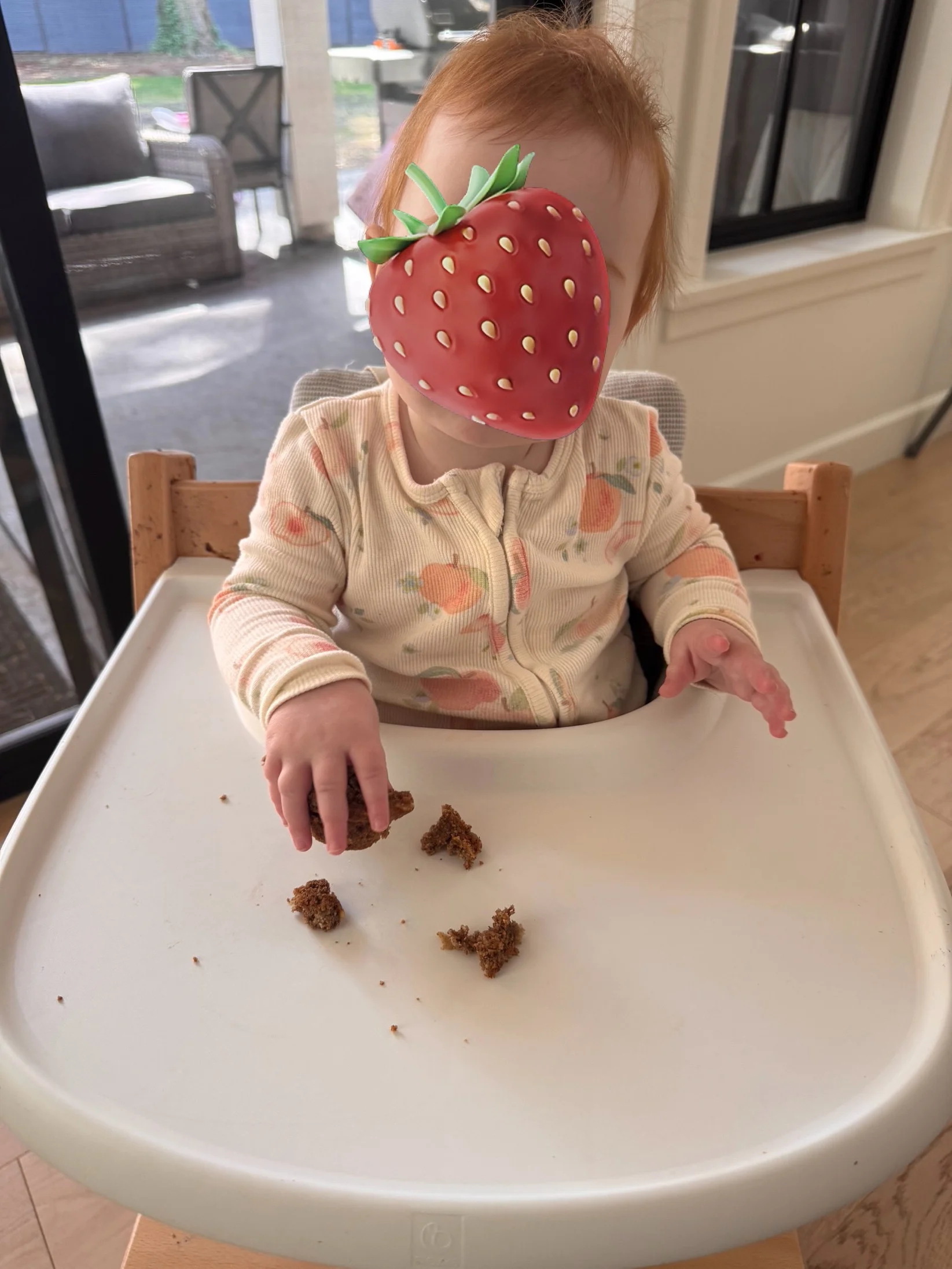 10 month baby with red hair sitting in a high chair, eating a prune muffin. Strawberry is blocking her face.